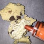 A cookie has been broken apart, and a white substance is being dispensed onto it from an orange tube, adding a surprise element. This could be part of a Cream Filled Chocolate Chip Cookies recipe, enhancing the treat's delectable appeal.