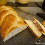 A loaf of braided challah bread with two slices cut, placed on a cutting board next to a knife. The background includes a blurred container and a yellow bottle, hinting at the traditional challah recipe used.