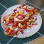 A plate of Challah French toast topped with diced strawberries, peaches, and a sprinkle of powdered sugar sits invitingly on a tiled table.