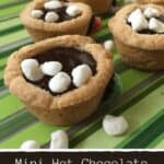 Four cookie cups topped with melted chocolate and small marshmallows are arranged on a green striped surface. The caption below reads: "Mini Hot Chocolate Cookie Cups Recipe" from cha-chingqueen.com.