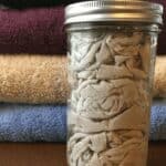 A mason jar filled with fabric scraps for DIY reusable fabric softener dryer sheets sits on a wooden surface in front of neatly folded towels in black, maroon, beige, and blue colors.