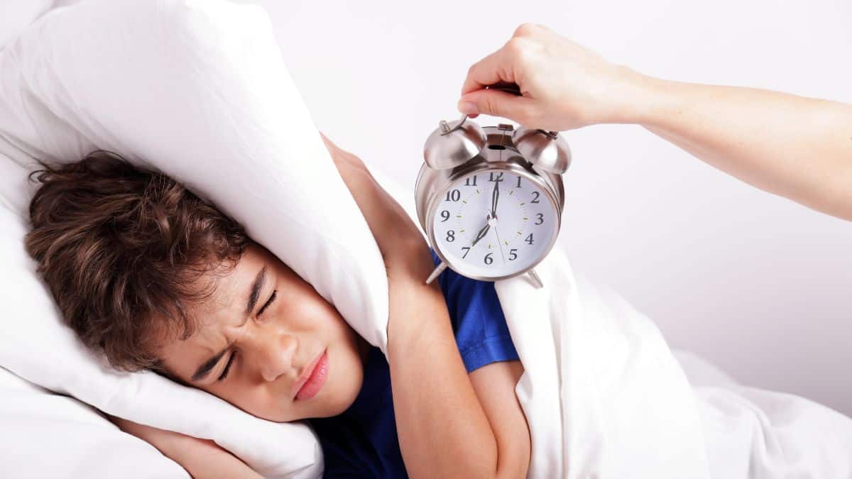 Tips For Waking Up Early; Teen Covering Ears with Pillow and Alarm Clock