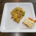 A square white plate with a serving of creamy pasta with broccoli and a slice of bread topped with melted cheese, set on a wooden table.