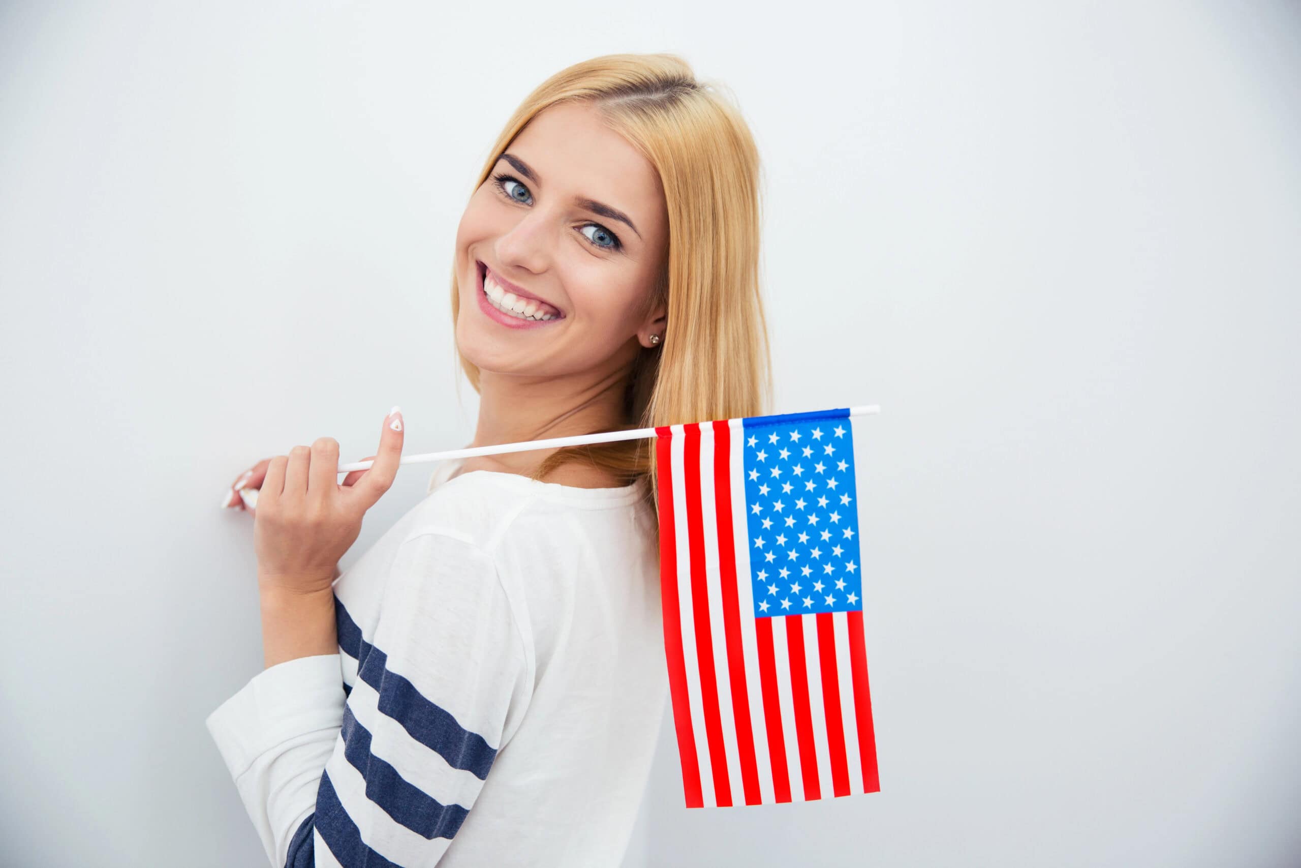 A smiling woman with blonde hair holds a small United States flag over her shoulder against a plain background.