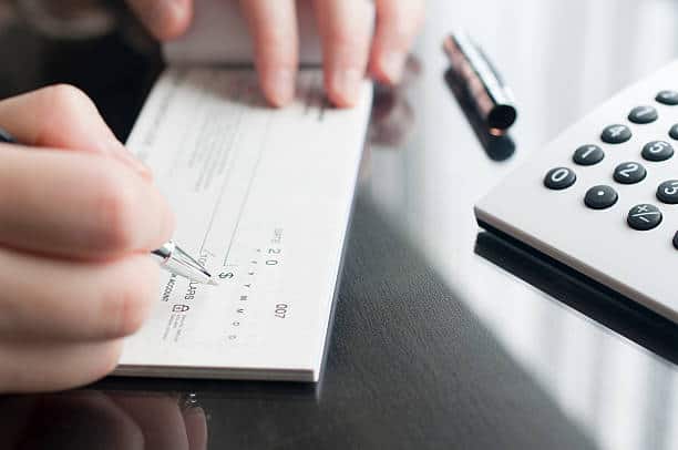 A person writing a check on a glass table with a pen, next to a calculator.