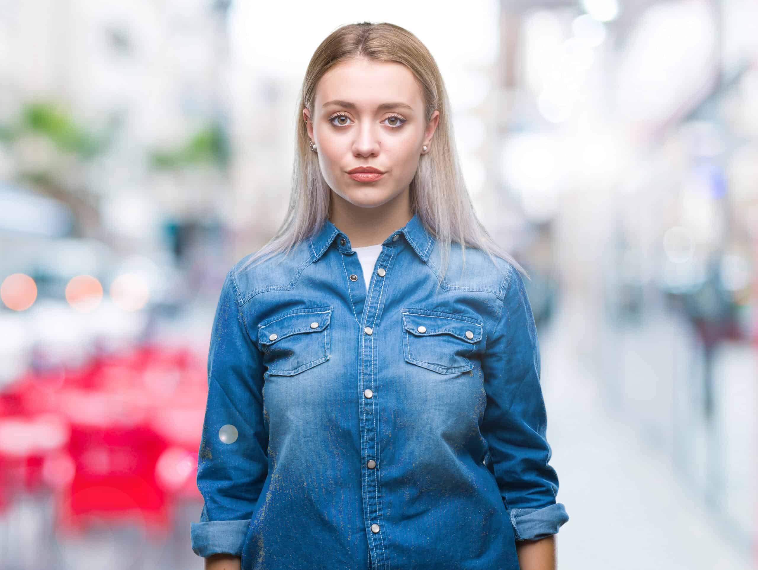 Woman with long blonde hair in a denim shirt standing outdoors on a blurred street with red chairs in the background.