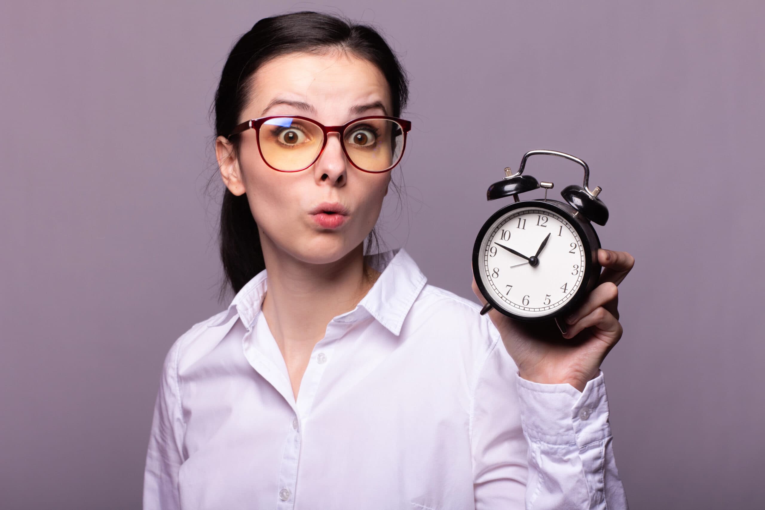 A woman in glasses and a white shirt holds a black alarm clock, looking surprised.