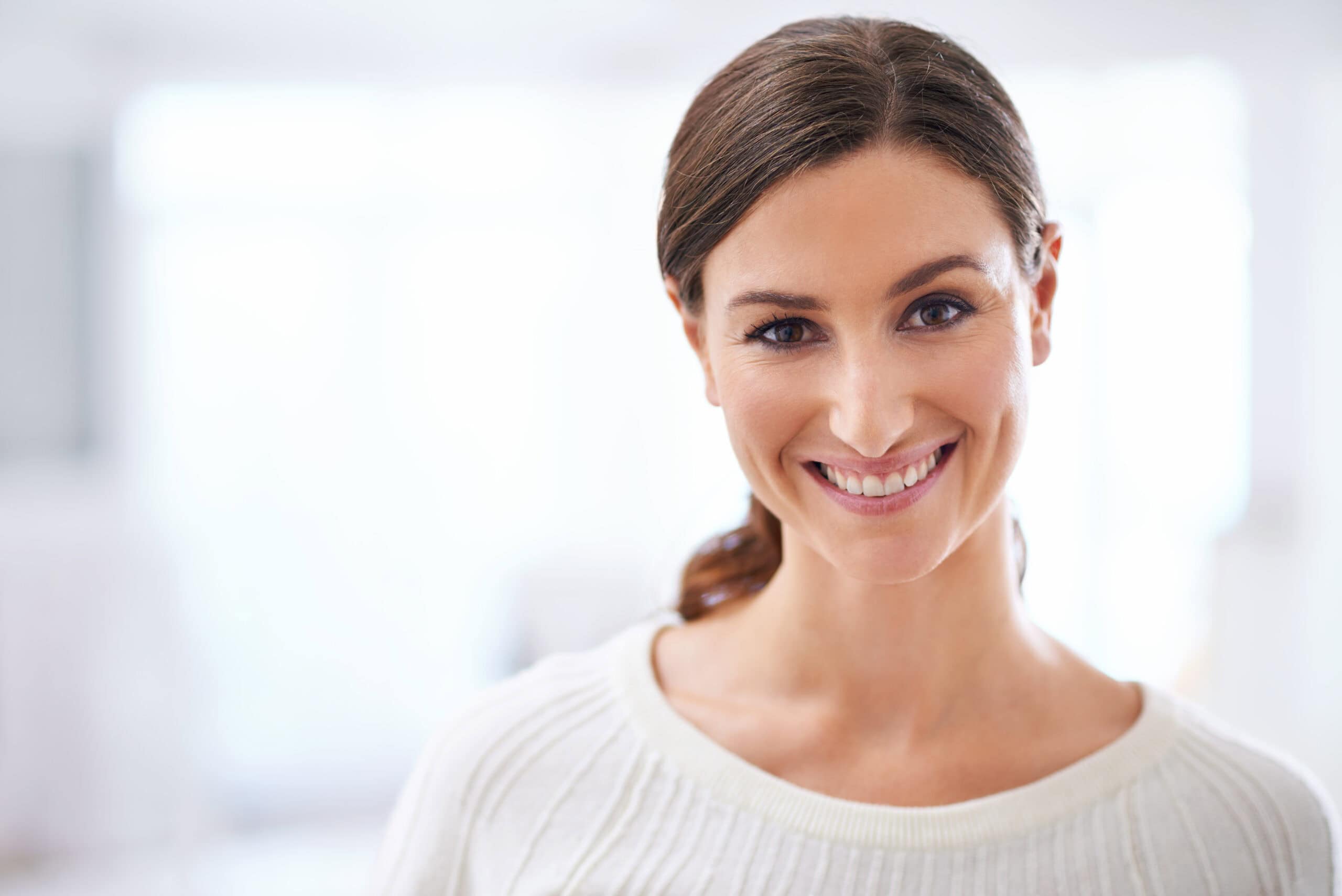 Smiling person with long brown hair in a white sweater, looking directly at the camera with a light blurred background.
