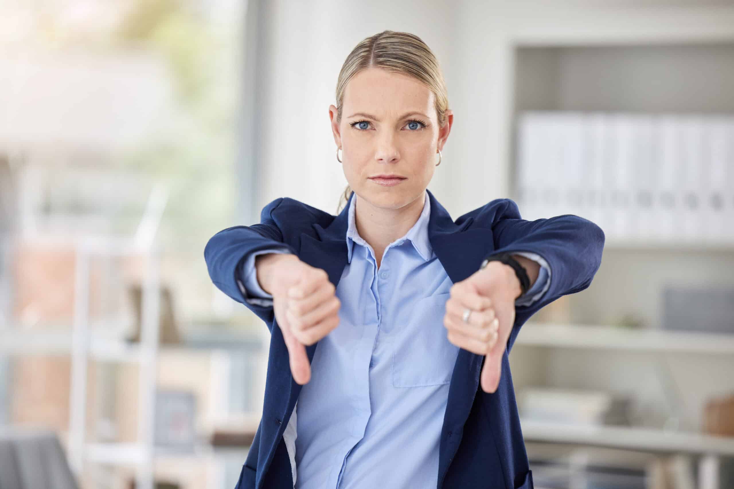 A woman in a blue suit with a serious expression gives two thumbs down in a bright office setting.