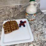 Waffle on a square white plate with blueberries and raspberries, next to a jug of organic maple syrup on a granite countertop.