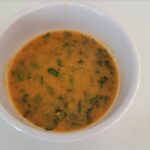 A white bowl filled with orange soup containing green herbs and vegetables.