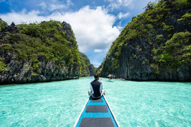 Person sitting at the front of a boat, surrounded by clear turquoise water and lush, rocky cliffs under a partly cloudy sky.