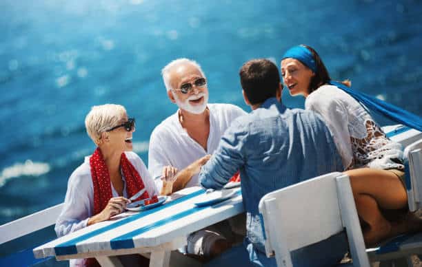 Four people sit at a table by the ocean, enjoying a sunny day. They are smiling and appear to be talking and eating.