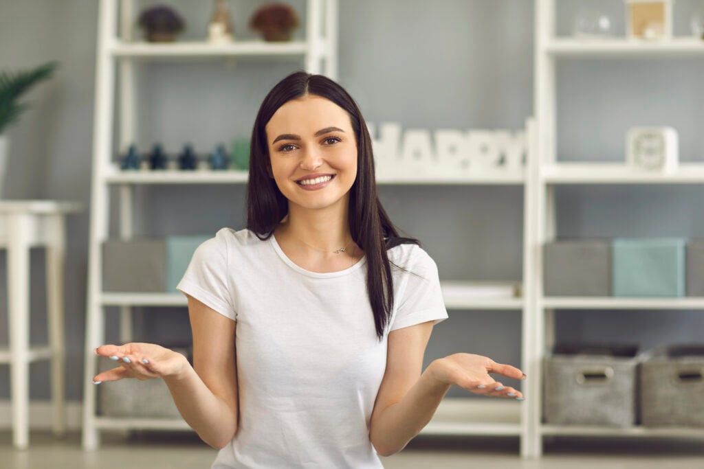 Woman with dark hair in a white t-shirt sits indoors, smiling and shrugging. Shelves with decorative items are in the background.