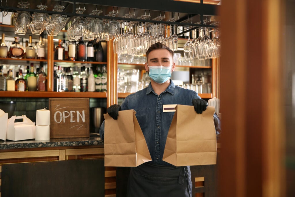 A man wearing a mask and gloves holds two paper bags in a restaurant with an "Open" sign behind him.