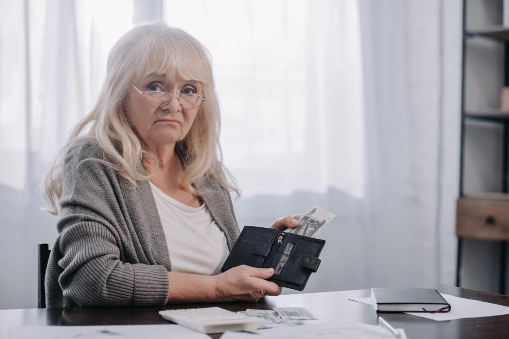 Elderly woman with long white hair, wearing glasses, holds a wallet with money, seated at a table with documents and a notebook.