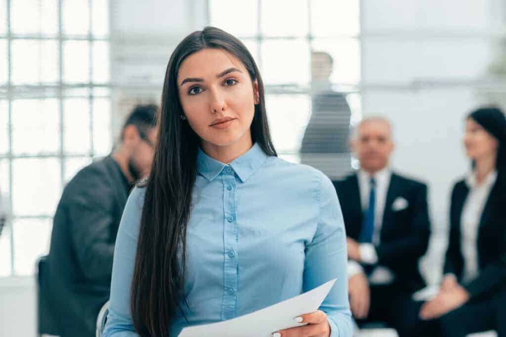 A woman in a blue shirt holds a document, standing in front of a blurred office meeting scene with four seated people.