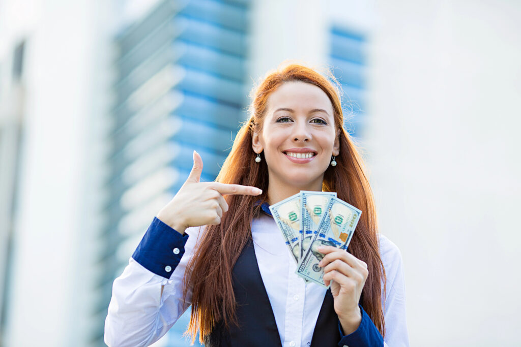 A woman with long red hair holds several US hundred-dollar bills in one hand and points to them with the other, smiling. She is wearing a white shirt and black vest.