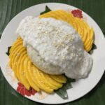 A plate of mango sticky rice with sliced mangoes, sticky rice, and coconut sauce topped with sesame seeds, placed on a banana leaf.