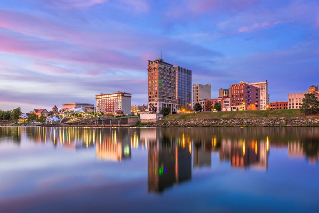 City skyline at dusk reflected on calm water, featuring multiple buildings, including a tall central building, with a colorful sky above.