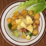 A bowl of fruit salad with mango, lettuce, tomatoes, onion, and apple slices on a wooden table.