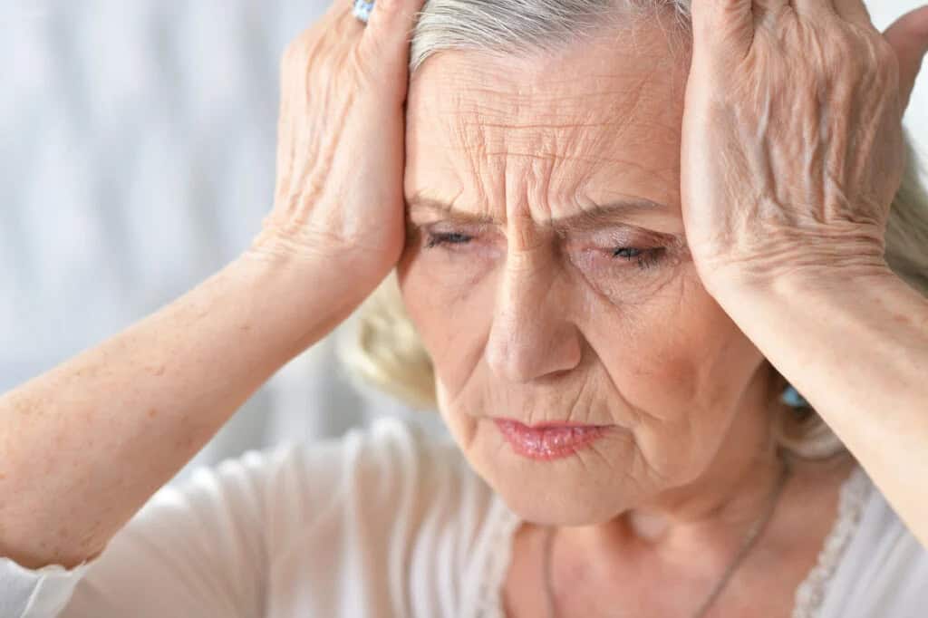 Elderly woman with gray hair holding her head with both hands, appearing to be in discomfort or deep thought.