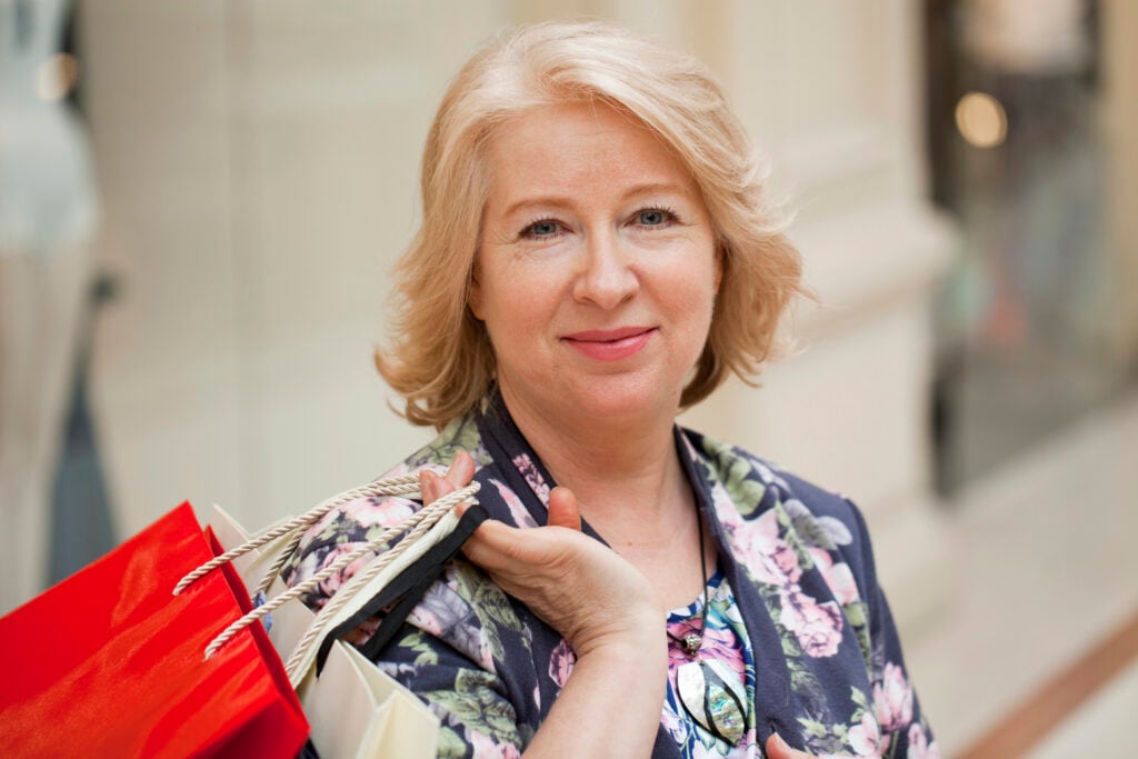 A middle-aged woman with light hair holds several shopping bags and smiles at the camera indoors.