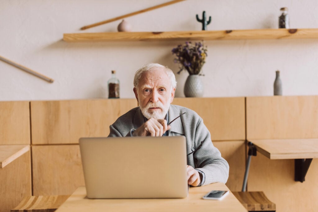 An elderly man sits at a table with a laptop, holding his glasses and looking at the camera; a smartphone is on the table and shelves with decor are in the background.