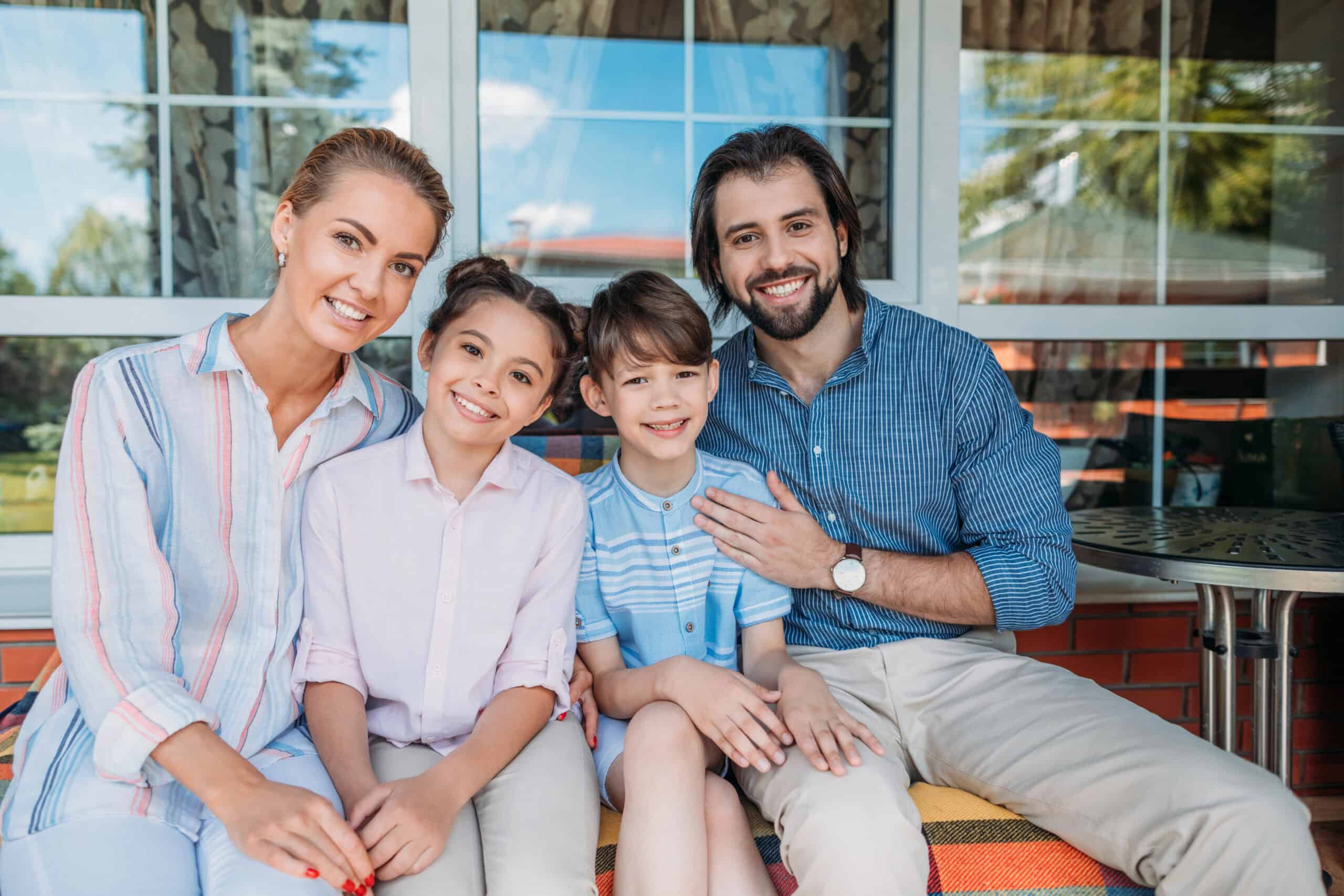 A family of four sitting on a porch. The parents are seated on either side of their two children. The background shows a window and part of a house. Everyone is smiling at the camera.