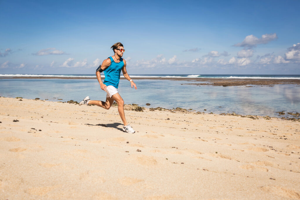 A man in a blue tank top and white shorts runs on a sandy beach near the ocean under a clear sky.