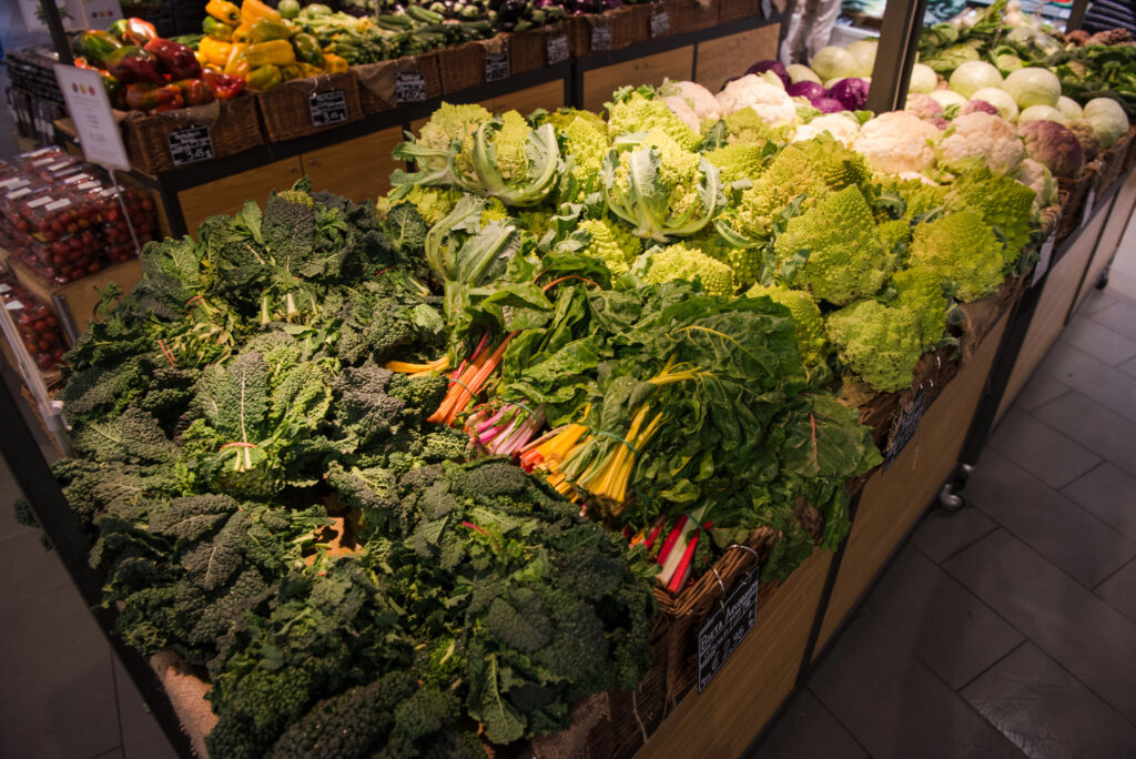 Assorted leafy greens and vegetables, including kale, romanesco, Swiss chard, and cauliflower, displayed on a grocery store produce table.