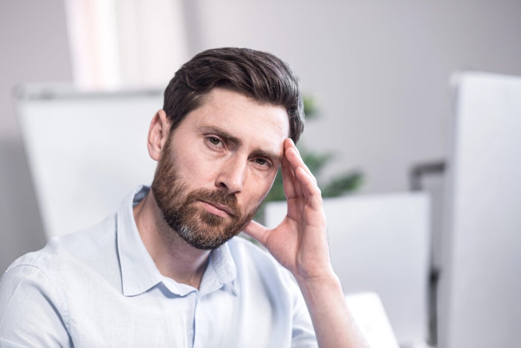 A man with a beard and light shirt sits indoors, resting his head on his hand and looking at the camera with a serious expression.