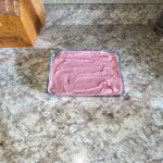 A glass dish on a marble countertop contains spreadable pink substance. A wooden knife block and a metallic container are in the background.