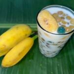 A glass of tapioca pearls with milk and fruit slices beside three ripe bananas on a green banana leaf.