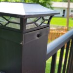 Close-up of a black outdoor solar post light mounted on a metal railing, with a green yard and wooden fence visible in the background.