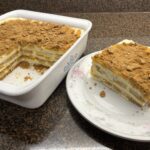 A layered dessert with cream and cookies, dusted with cocoa powder, is shown in a baking dish with one square piece served on a plate.