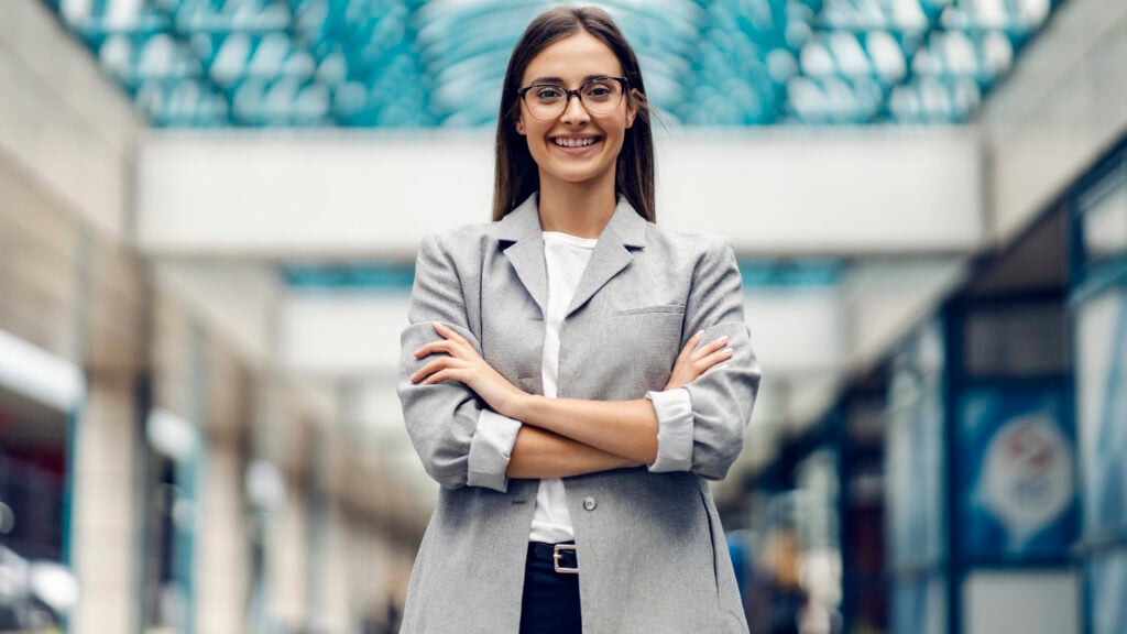 A person wearing glasses and a gray blazer stands indoors with arms crossed, smiling at the camera. The background features a glass ceiling and blurred surroundings.