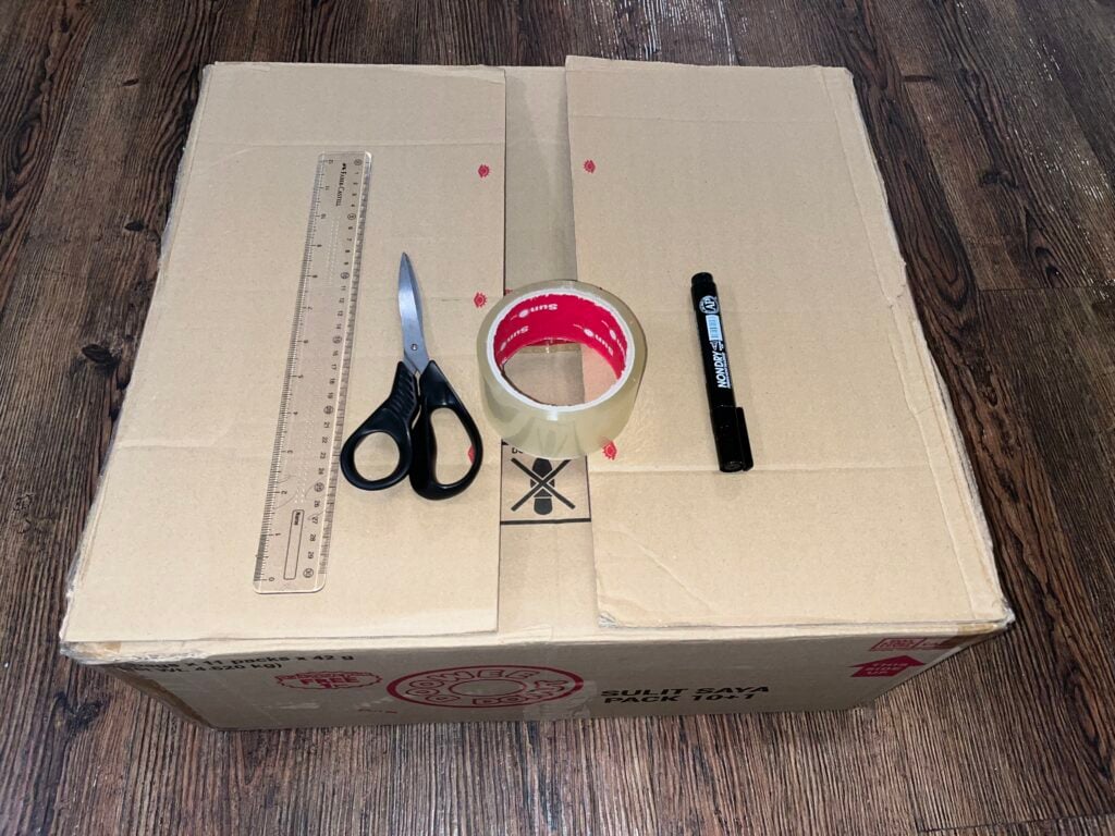 A closed cardboard box on a wooden floor with a ruler, scissors, a roll of clear tape, and a black marker placed on top.