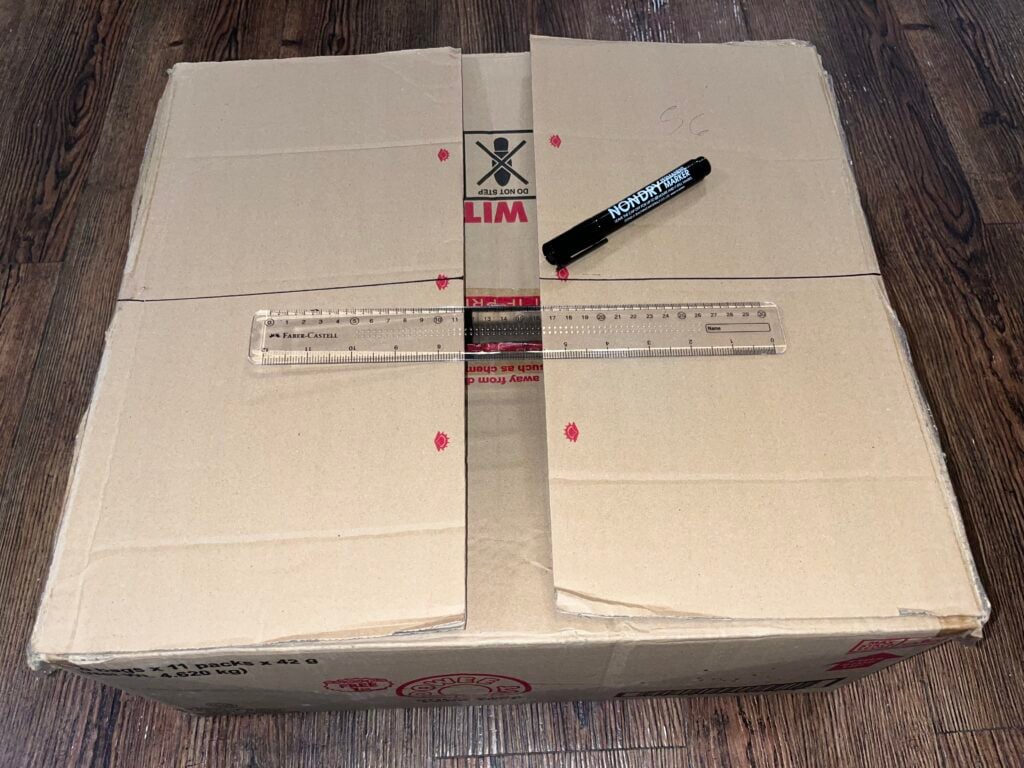A cardboard box with four flaps partially cut, a clear plastic ruler, and a black marker lying on top, placed on a wooden floor.