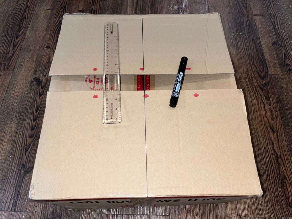 A closed cardboard box on a wooden floor with a clear ruler and a black marker placed on top. Red dot markings and a blue line are drawn on the box flaps.