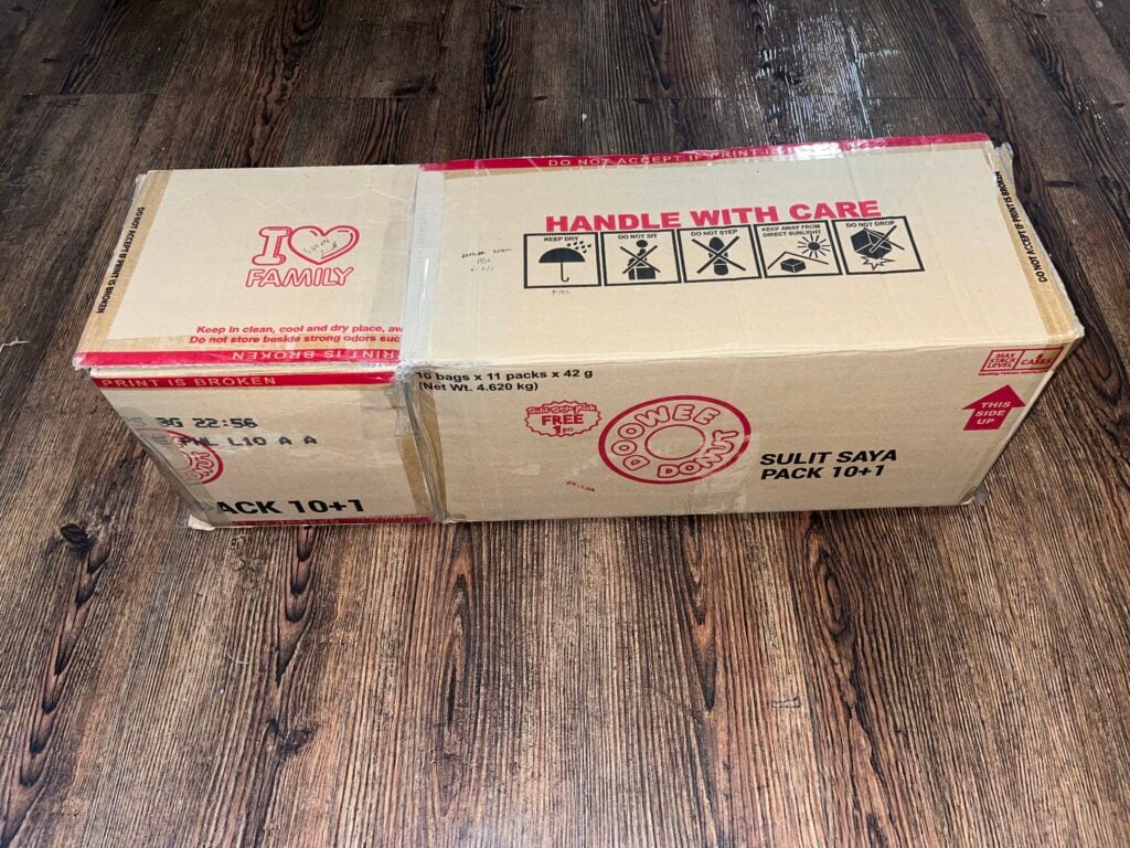 A closed cardboard box with red text and symbols, labeled "HANDLE WITH CARE" and "GOODIE DOS", sits on a wooden floor.
