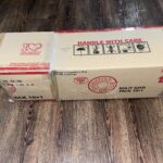 A closed cardboard box with red text and symbols, labeled "HANDLE WITH CARE" and "GOODIE DOS", sits on a wooden floor.