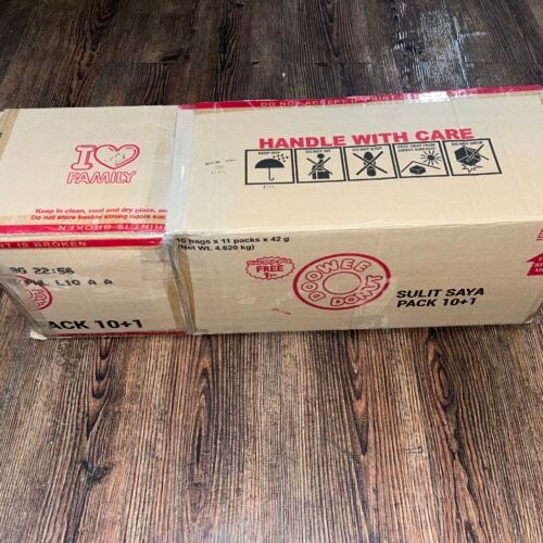 A closed cardboard box with red text and symbols, labeled "HANDLE WITH CARE" and "GOODIE DOS", sits on a wooden floor.