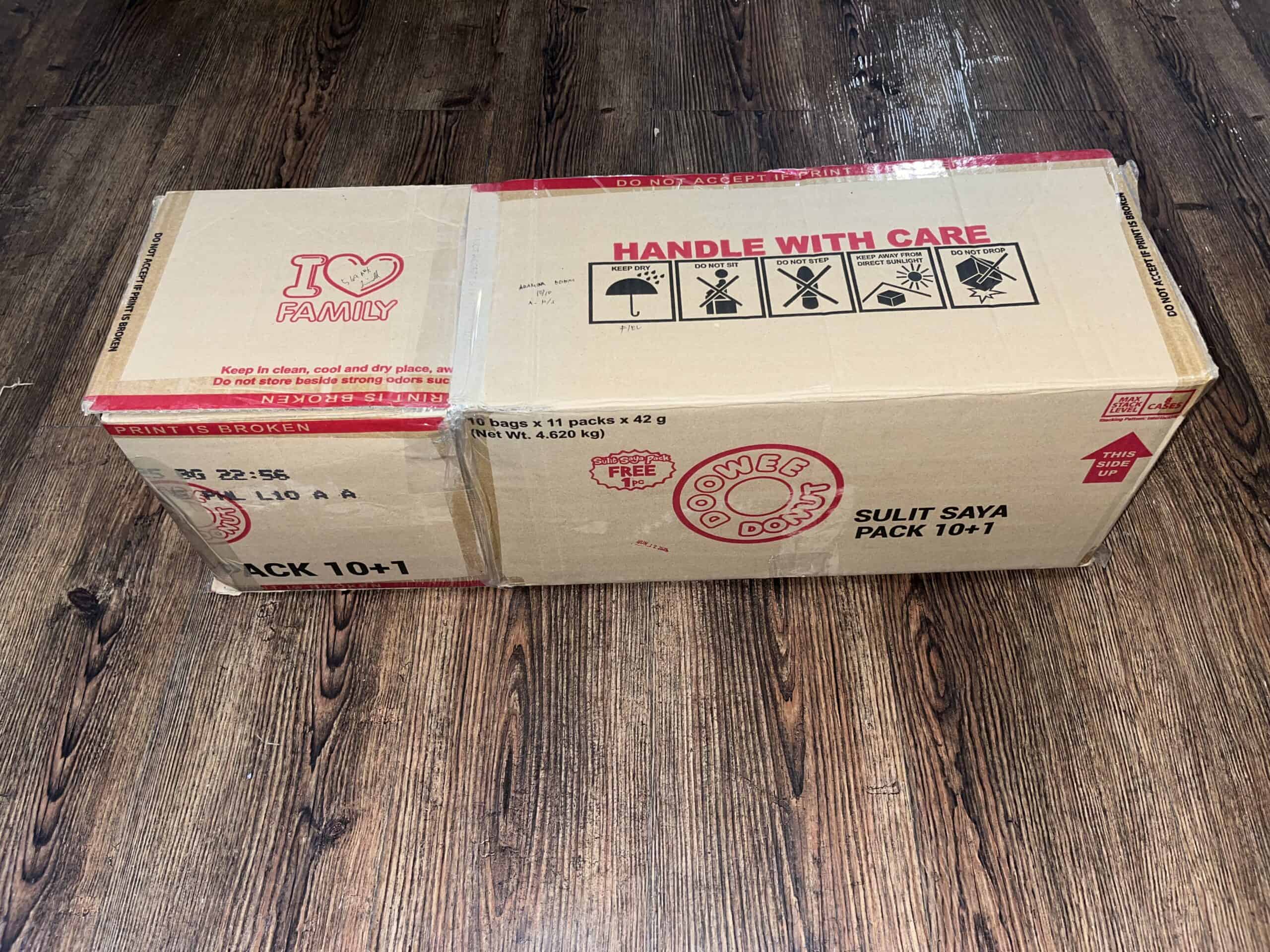 A closed cardboard box with red text and symbols, labeled "HANDLE WITH CARE" and "GOODIE DOS", sits on a wooden floor.