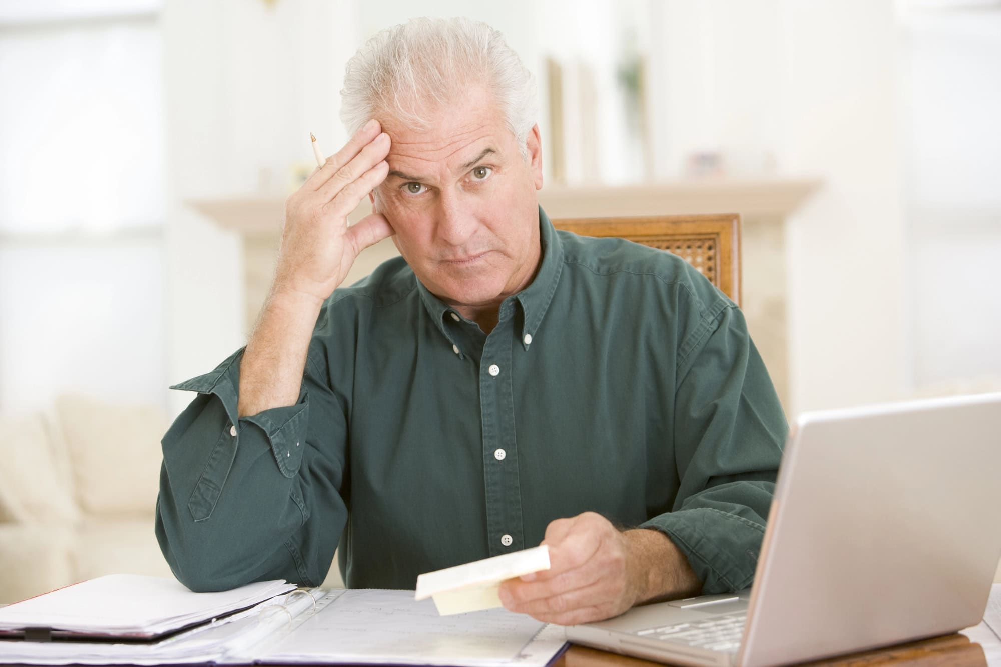 Older man in a green shirt sitting at a table with a laptop, holding a bill or receipt, looking concerned or stressed.