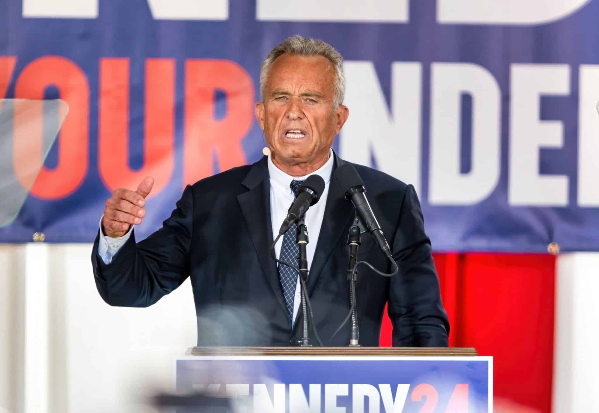 A man in a suit speaks emphatically at a podium with multiple microphones; a campaign sign reading "KENNEDY 24" is visible behind him.