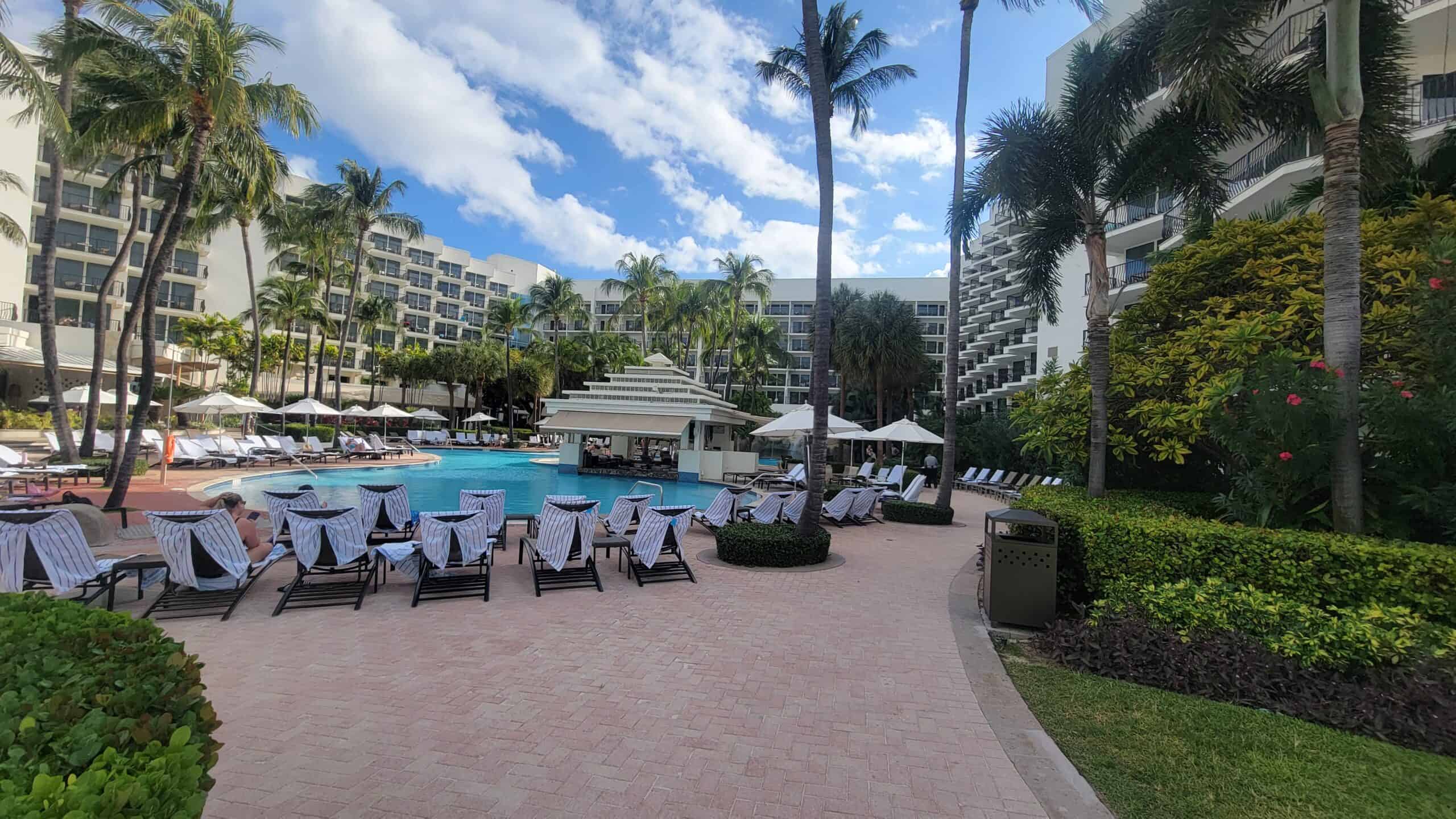Outdoor hotel pool area at Aruba Marriott Resort with lounge chairs, umbrellas, and palm trees, surrounded by multi-story buildings under a partly cloudy sky—perfect for an anniversary getaway or visiting Stellaris Casino nearby.