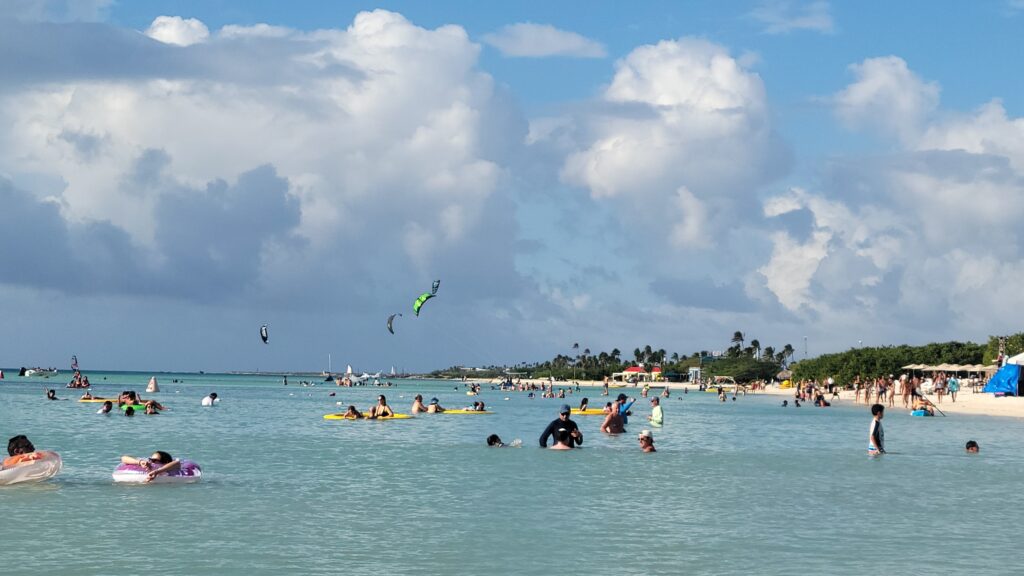 People swimming, relaxing on floats, and windsurfing in a shallow, crowded beach area under partly cloudy skies with palm trees and Aruba Marriott Resort’s Stellaris Casino in the background—perfect for an anniversary getaway.