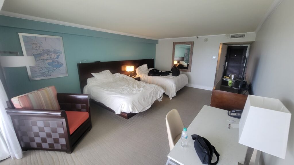 Hotel room at the Aruba Marriott Resort features two unmade beds, a desk and chair, armchair, dresser with mirror, and a lamp—perfect for an anniversary getaway near Stellaris Casino. Personal items rest on the beds, desk, and dresser.