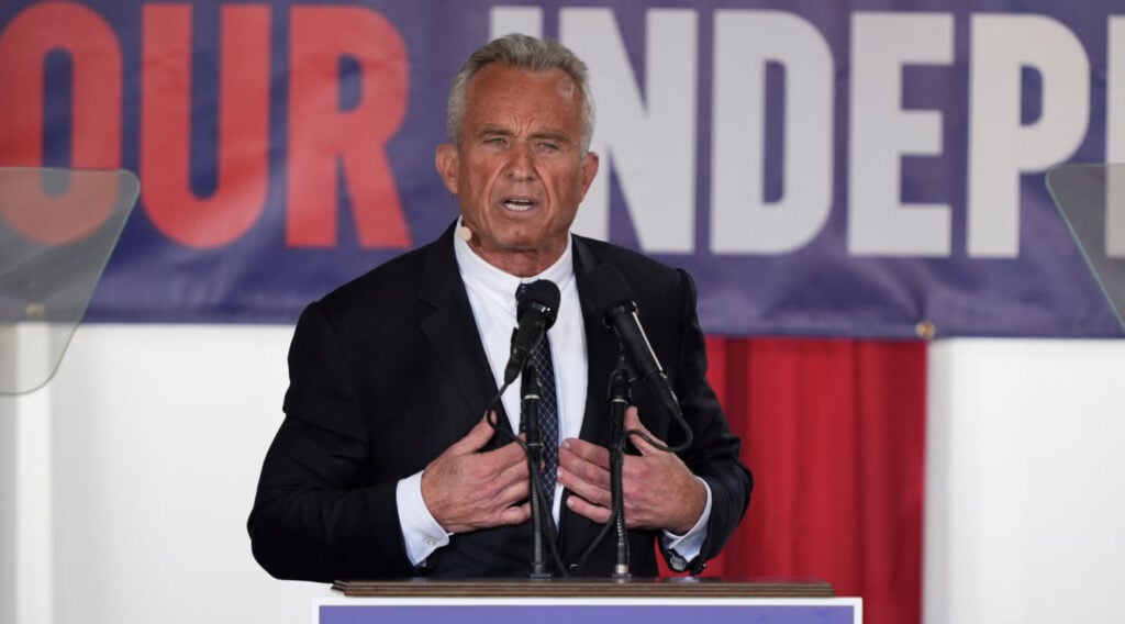 A man in a suit speaks at a podium with two microphones, standing in front of a banner that partially reads "INDEPEN.